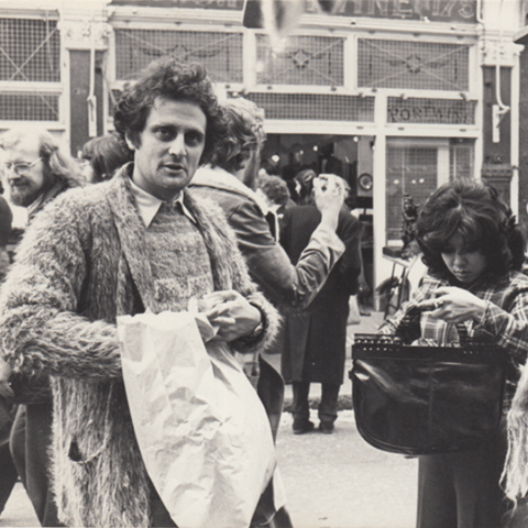 A sepia-toned image of a man in a market, looking at the camera while holding a bag