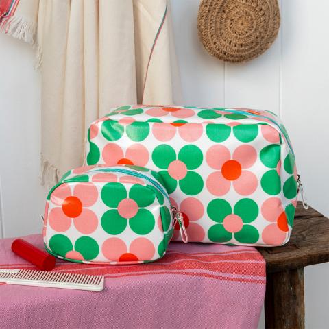 A wash bag and a makeup bag with bright green and pink floral print sit on a bench with a comb and lipstick