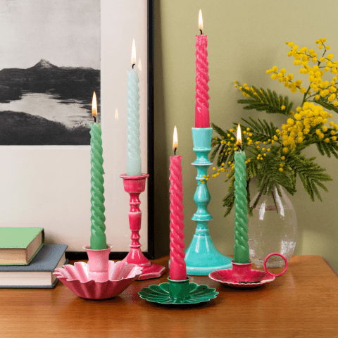 Colourful candlesticks with candles sit on a brown desk with a plant and books in the background