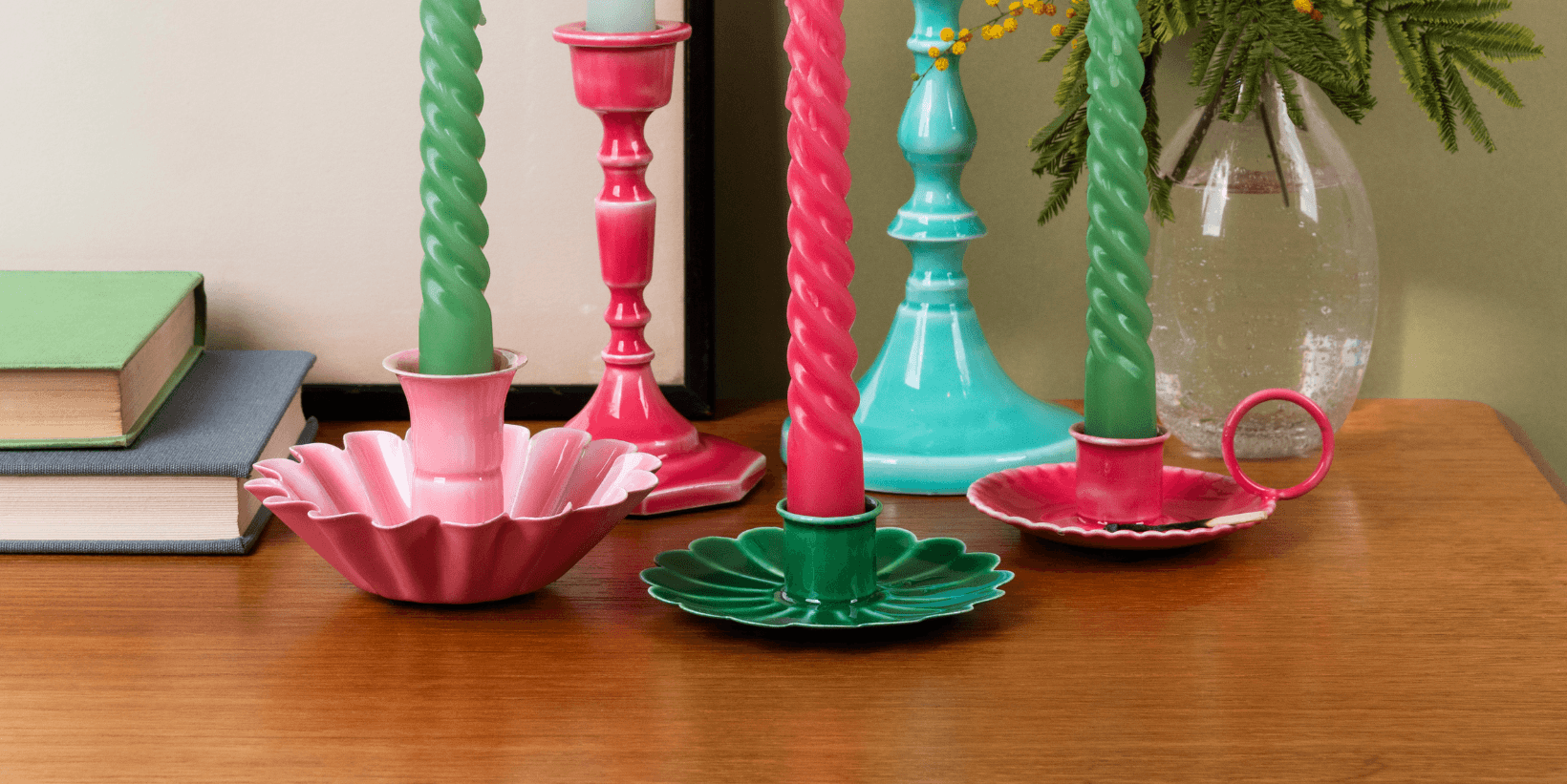 Colourful candlesticks with candles sit on a brown desk with a plant and books in the background