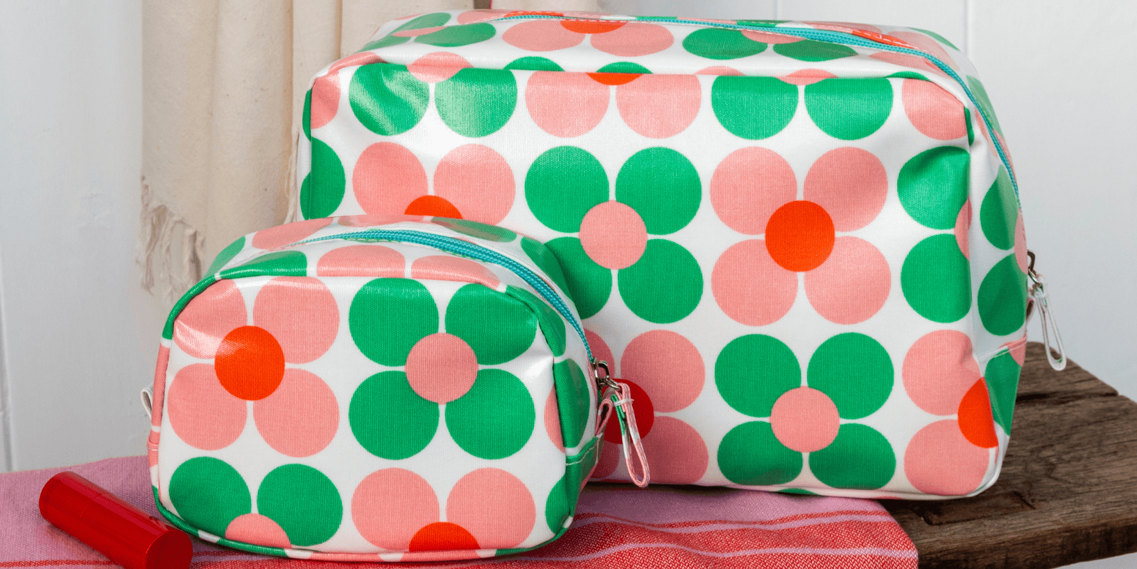 A wash bag and a makeup bag with bright green and pink floral print sit on a bench with a comb and lipstick