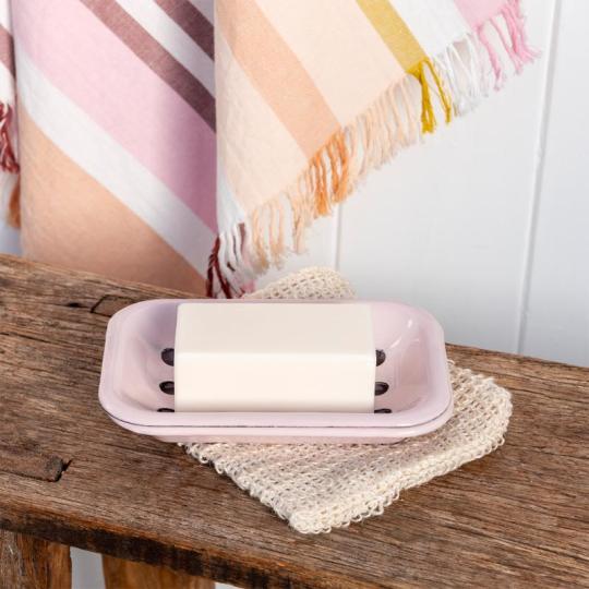 A pale pink soap dish sits on a wooden bench with a bar of soap inside