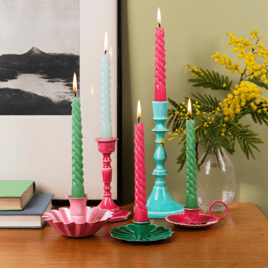 Colourful candlesticks with candles sit on a brown desk with a plant and books in the background